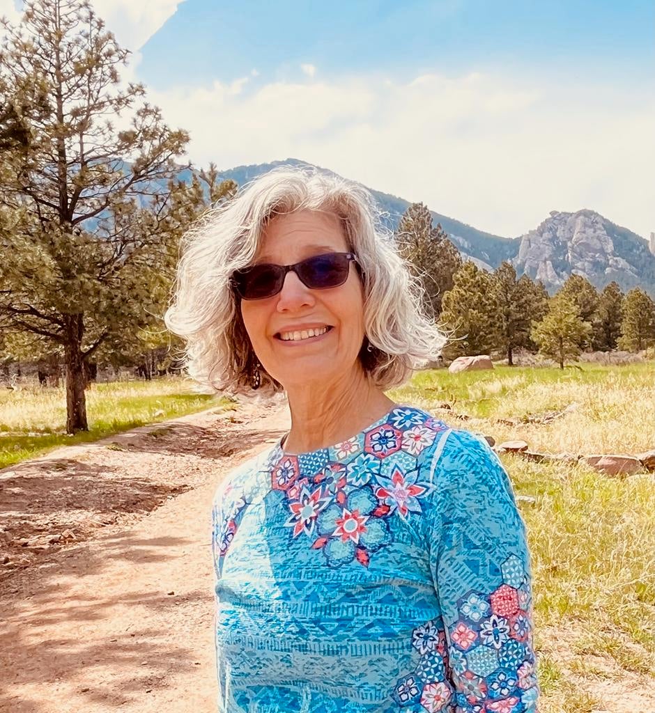 Carolyn A. Wilson, Ph.D. stands on a trail in front of a mountain range, wearing a bright blue shirt.