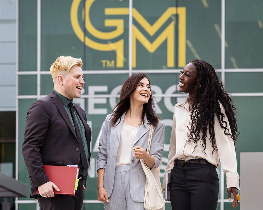 Three George Mason students wearing business suits stand together in front of Mason Square. 