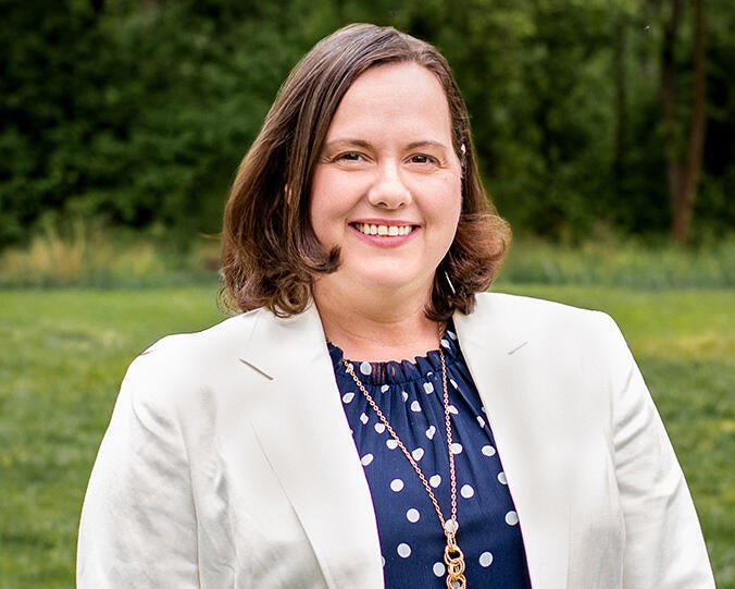 Danielle Passano is wearing a cream colored blazer over a blue and white polka-dot blouse, she is outside in a green field smiling at the camera.