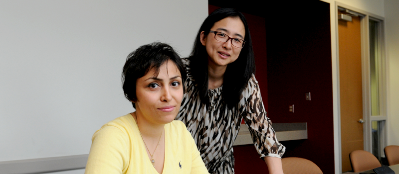 Two women in casual attire smiling at camera - one wearing yellow top, other in patterned dress with glasses