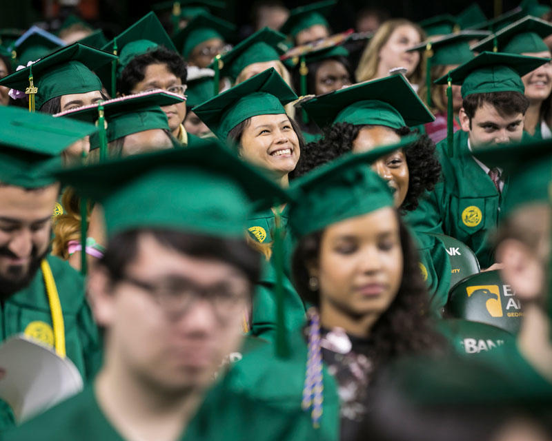 Crowd of graduates in regalia, one person at the center of the crowd looking up, smiling hopefully. 