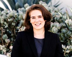 Nina Cox has shoulder length brown hair and she's wearing a dark blazer over a navy shirt and standing in front of a floral display